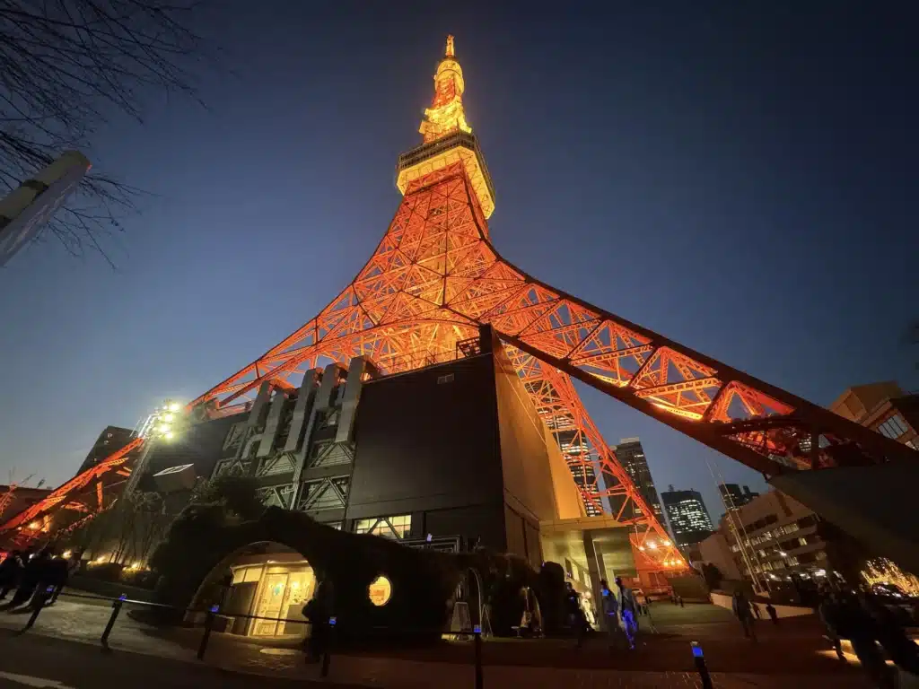 廣角仰拍東京鐵塔底部,塔身在深藍色夜空中散發強烈橘光,下方為 foot town 建築體。