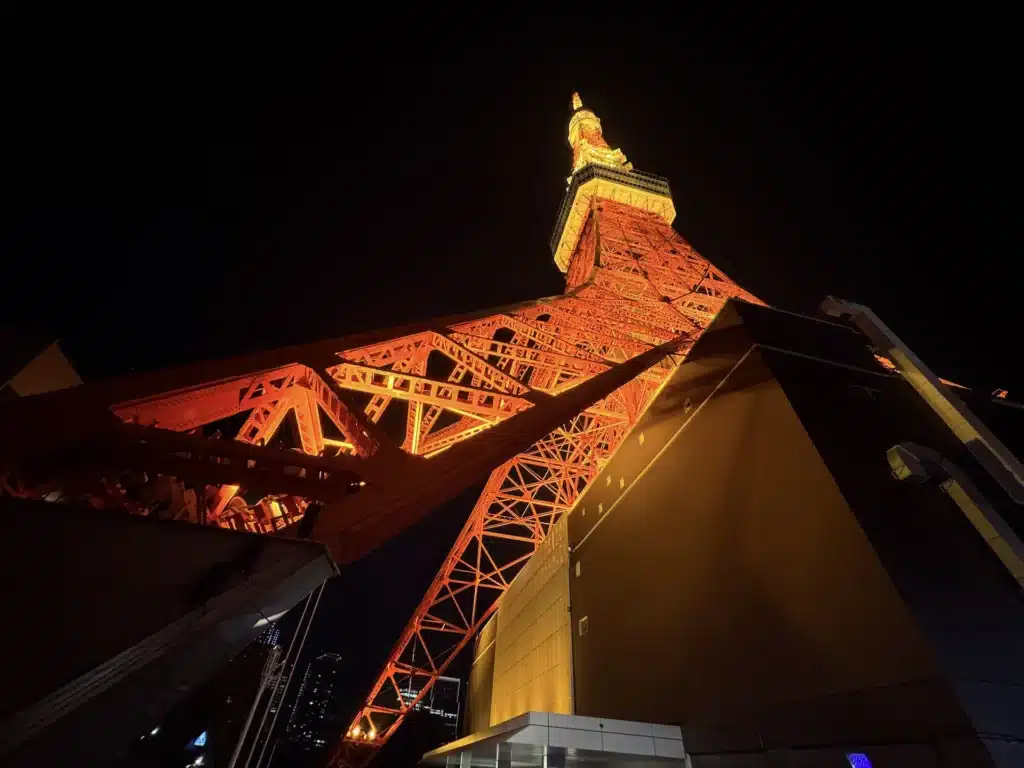 夜間拍攝東京鐵塔的斜側視角,深色的建築外牆與明亮的橘紅鋼架形成明暗對比。