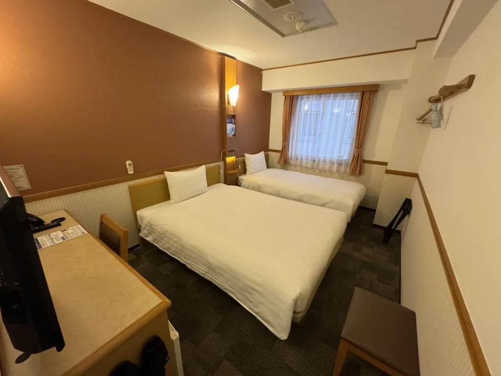 東橫 INN(高崎站西口)雙床房,兩張白色床鋪、書桌與窗邊的簡單小空間。