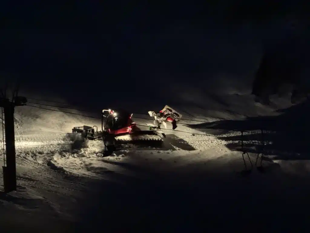 夜間的滑雪場中,一台紅色整地車近距離在推雪並整理坡面,車燈照亮前方雪塵飄散。
