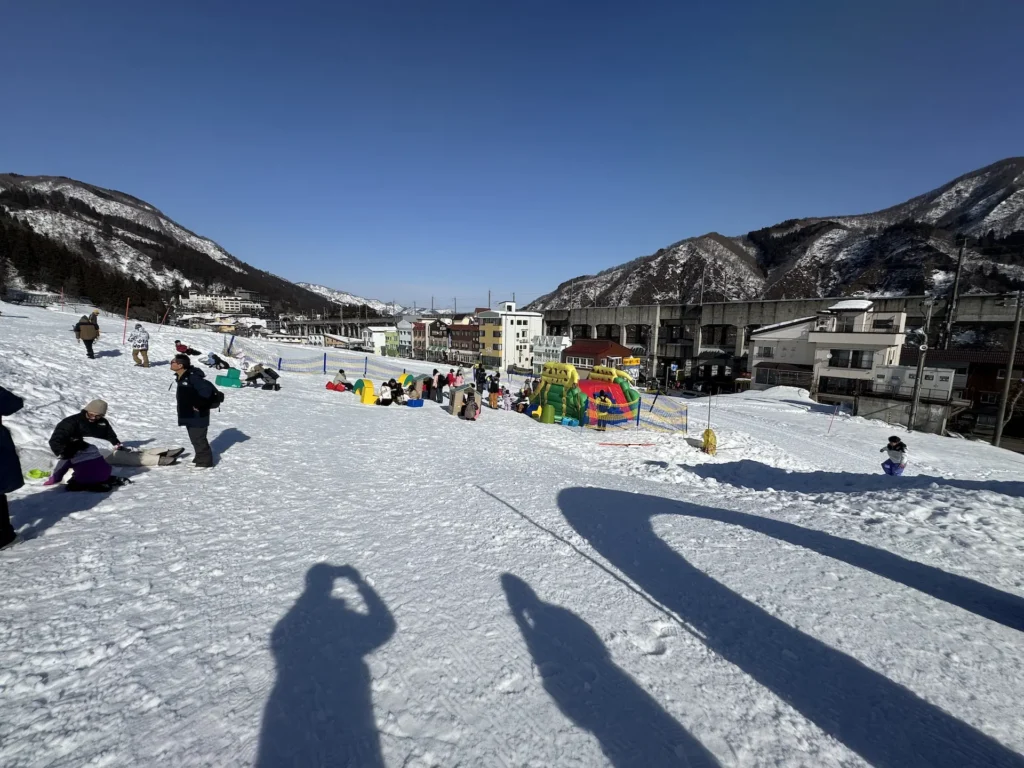 湯澤中里雪地遊戲區,全景可見家長與小孩在雪地上玩耍,背景是群山與住宅建築。