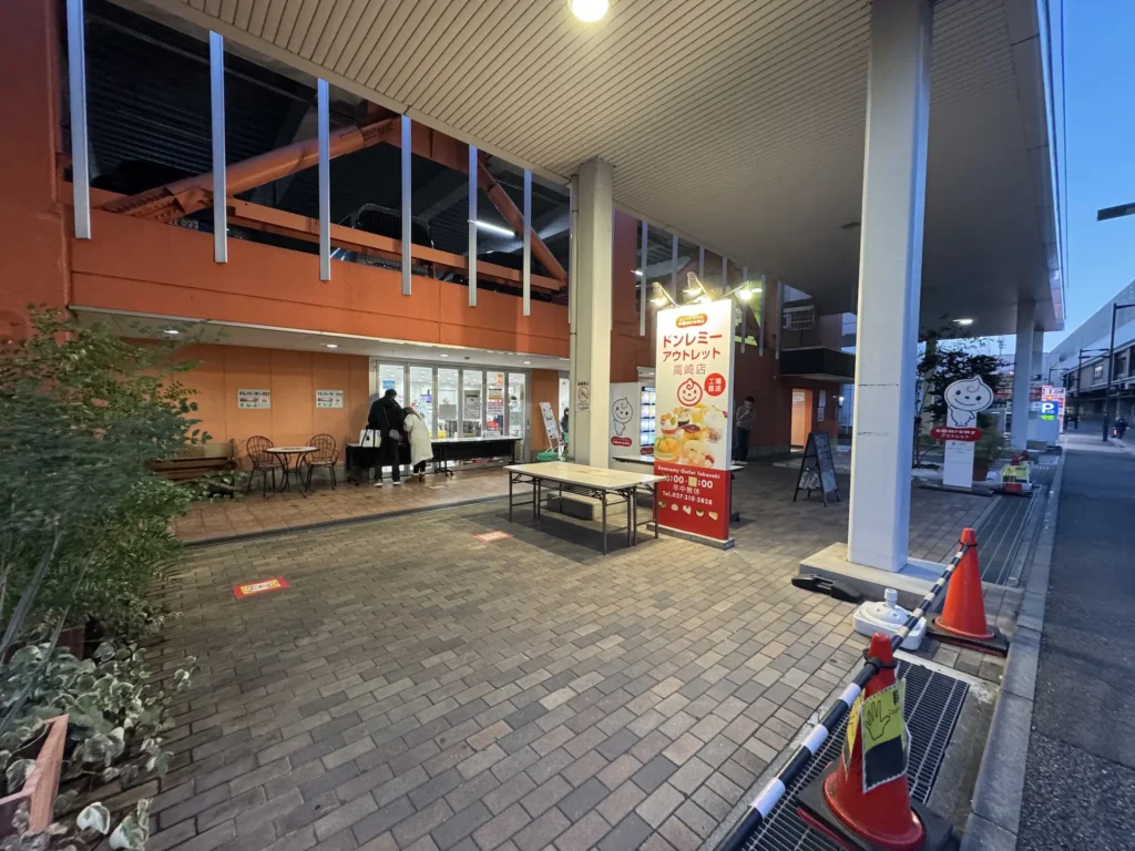 傍晚時分的 Domremy Outlet(ドンレミーアウトレット)高崎店外觀,入口前有紅色立牌和簡單座位區。
