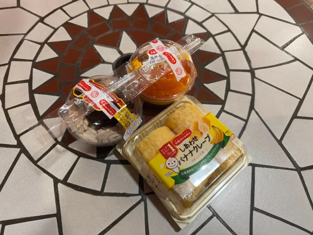 三款從 Domremy Outlet 購買的甜點,包括巧克力布丁、橘子果凍與香蕉可麗餅,擺放在花磚桌面上。