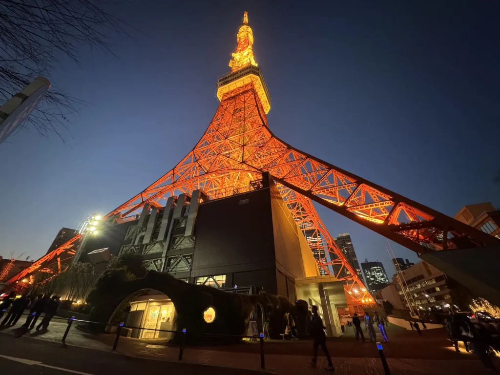 夜晚近距離仰拍東京鐵塔,塔身燈光點亮,遊客在下方拍照