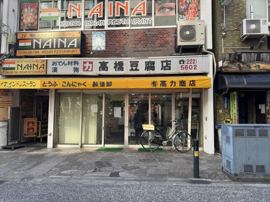 日本街邊的高橋豆腐店外觀,黃色遮雨棚,上方寫著「豆腐・こんにゃく製造卸」,門口停著一台腳踏車。