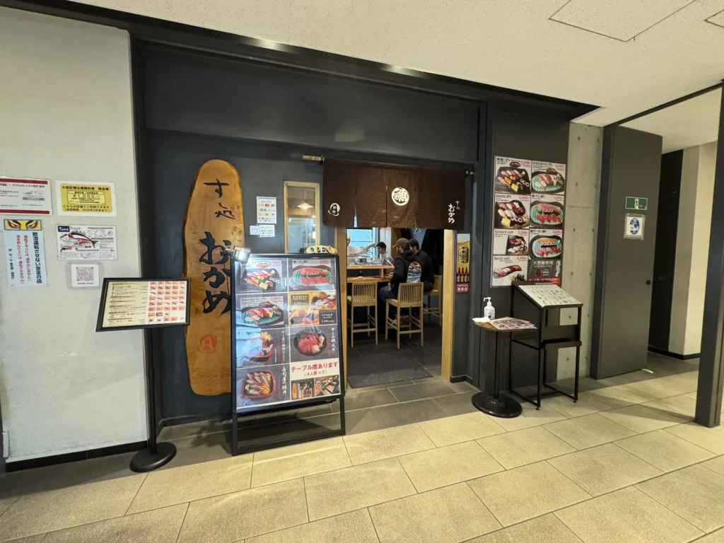 すし処おかめ的店面外觀,木製門面、布簾與擺在門口的握壽司照片看板。