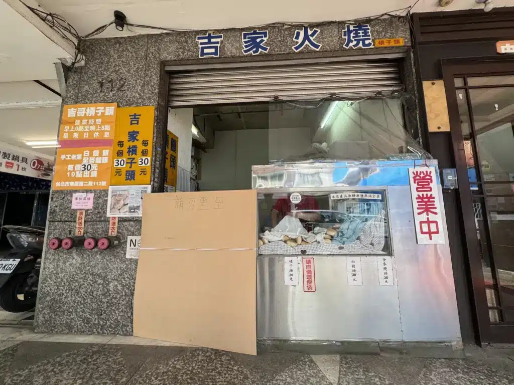 台北市南昌路二段「吉家火燒（槓子頭）」的實體店面全景。店面外掛有深藍色招牌，寫著「吉家火燒」與「槓子頭」。門口擺放著鮮黃色的廣告看板，詳細標註營業時間為早上9點至晚上8點、星期日休息，以及每個槓子頭、白饅頭和全麥饅頭皆為30元的價格，並註明「10點出鍋」。右側玻璃櫥窗上貼有「營業中」的紅字告示，內部可見擺放的槓子頭。