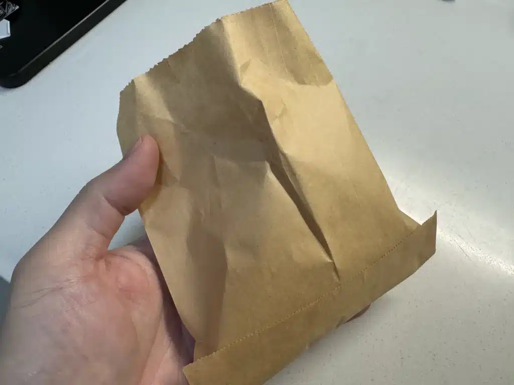 一隻手拿著一個裝有食物的棕色紙袋。紙袋底部有明顯的鋸齒狀邊緣和折痕，背景為一個乾淨的白色光滑桌面。