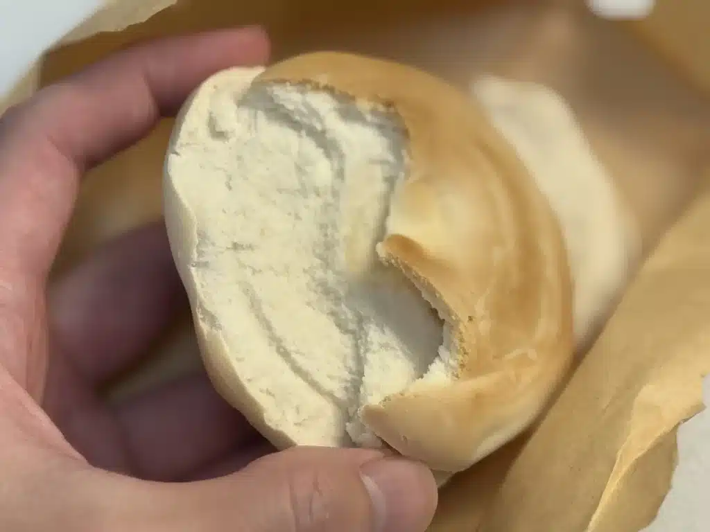 一隻手將一個槓子頭掰開，露出內部雪白、紮實且幾乎沒有氣孔的麵團組織。槓子頭焦脆的外皮和內部緻密的麵心形成鮮明對比，背景是撕開的棕色紙袋。