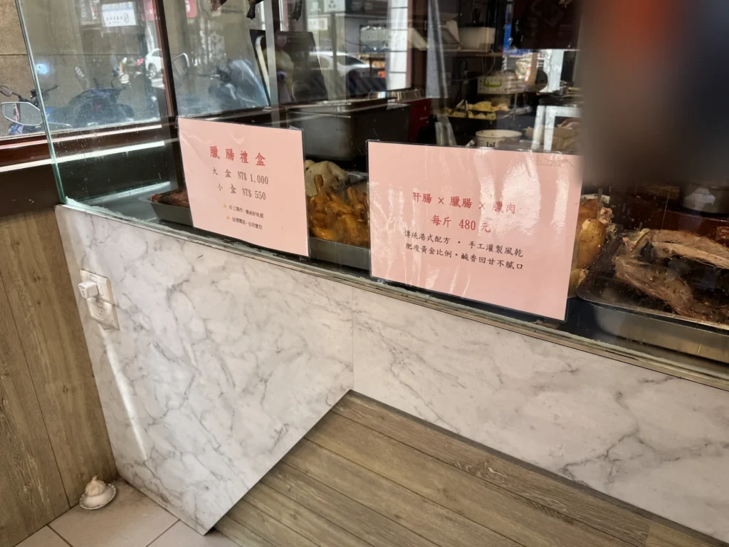 1+1 燒臘店門口的玻璃櫃，貼著臘腸禮盒與肝腸臘肉的價格